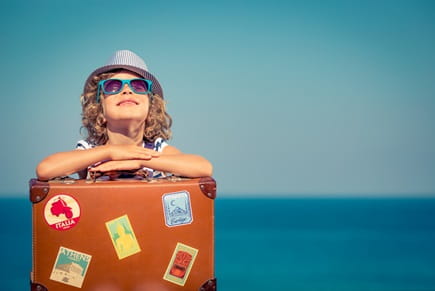 A little girl, wearing a hat and sunglasses, sits with her suitcase in front of the ocean