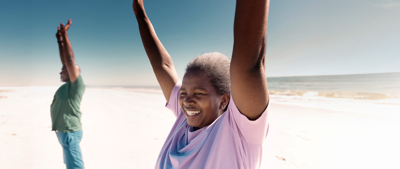 An elderly couple standing on the beach with their arms stretched above their heads.
