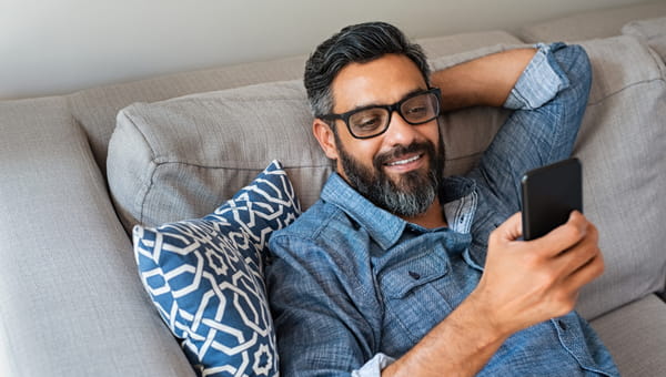 Happy smiling latin man using smartphone device while sitting on sofa at home