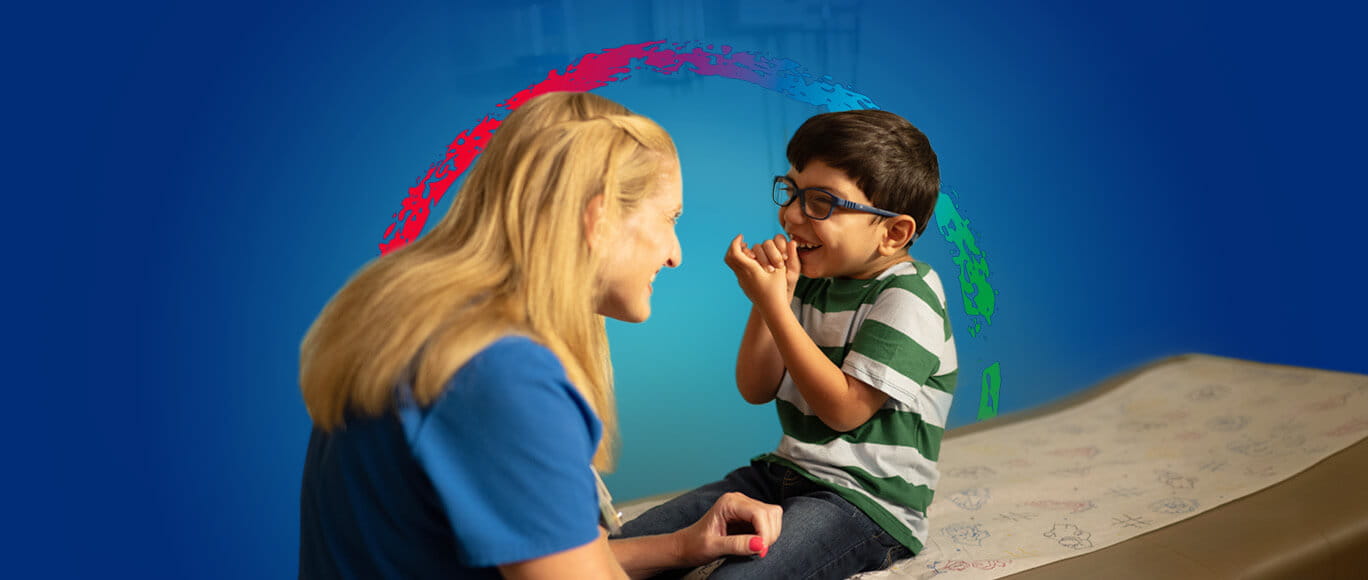 A young boy with glasses sitting on an examination bed, laughing with a pediatric specialist in a medical setting. They are surrounded by the BayCare Kids logo.