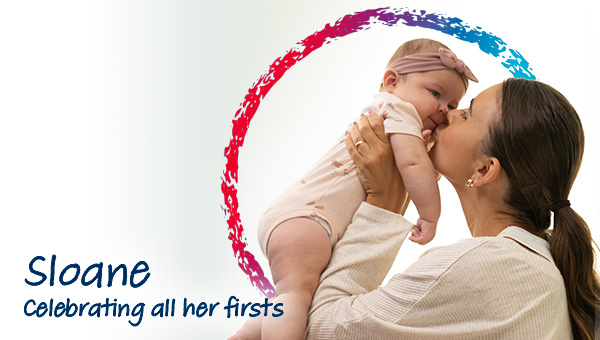 A woman embracing a 4-month-old baby, lifting them up and kissing their cheek lovingly. The photo has words that say, Sloane, celebrating all her firsts.