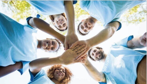 A group of men and women are standing in a circle and have extended their right arms so they can place their right hands on top of each other.