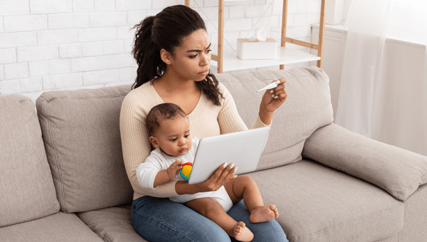 young mother holding newborn on couch and checking thermometer