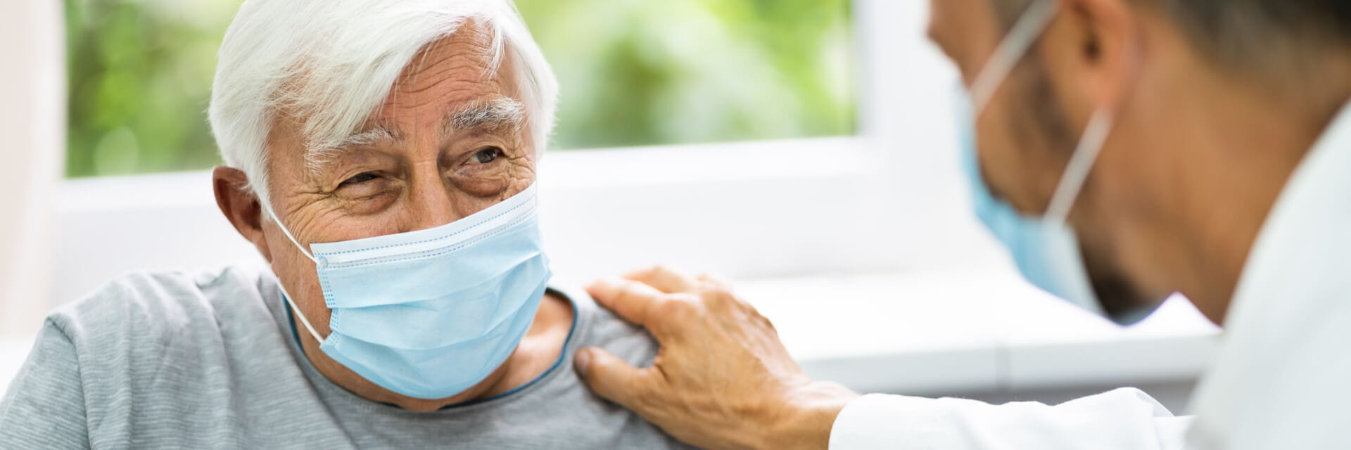 A senior man, who is wearing a mask, is visiting his doctor, who is also wearing a mask.