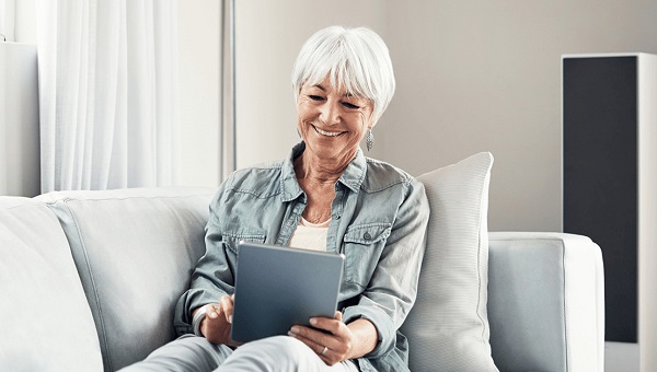 A senior woman is sitting on her sofa while looking at information on her tablet.