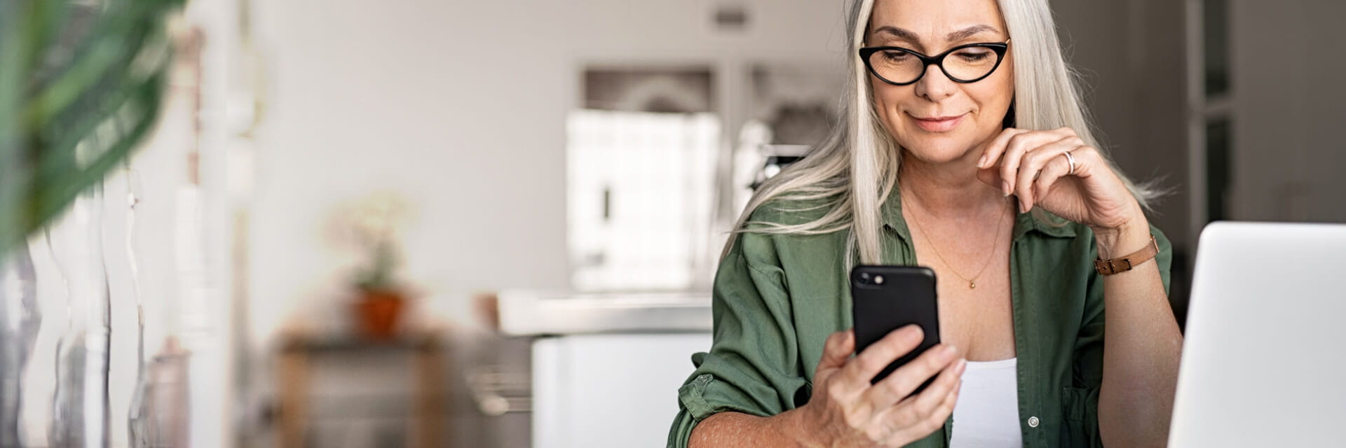 mature female looking at a mobile phone while sitting at a desk with a laptop