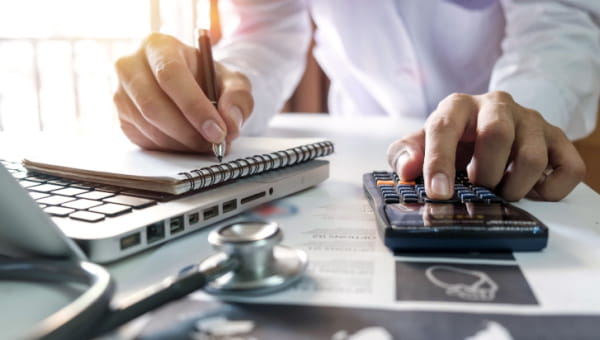 Male hand using a calculator and other hand taking notes in a notepad with a laptop and stethoscope on a desk.