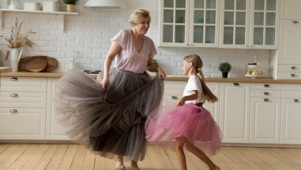 A grandma and grand daughter wearing tutus dance in the kitchen.