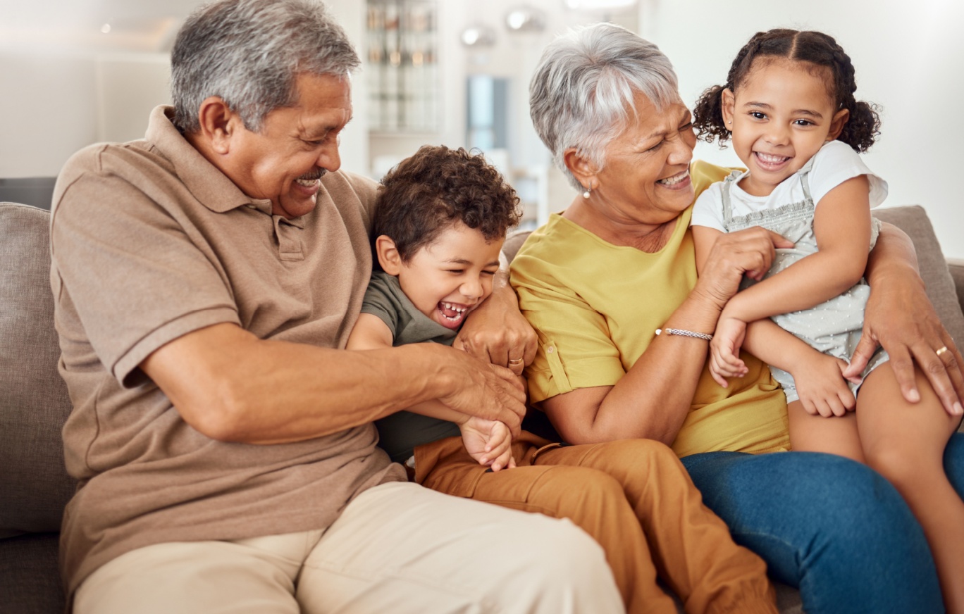 A happy family with grandparents tickling their grandchildren on a sofa.