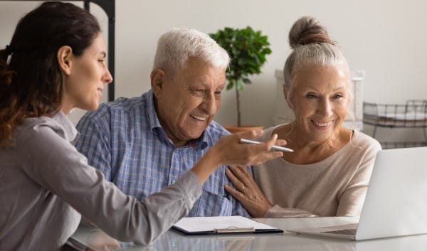 An elderly couple smiles while a younger woman points at something on the laptop in front of them.