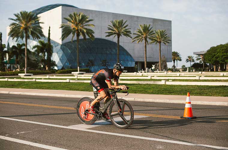 Male athlete riding bicycle and wearing athletic outfit riding on street in front of large museum with palm trees in front.