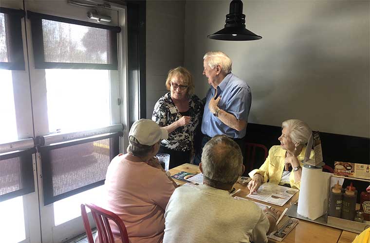 Woman standing next to man in blue shirt with gray hairs smiles and woman seated wearing a pink shirt and baseball cap. A woman with gray hair wearing a yellow jacket is smiling. A man is seated with his back to the camera. They are at a restaurant.