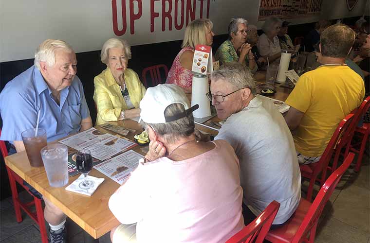 A gray-haired man in a blue shirt sits next to a gray-haired woman in a yellow jacket. They are across from a woman in a pink shirt and a man with glasses. They are talking and looking at menus at a restaurant. Other people are also at the table talking to each other.