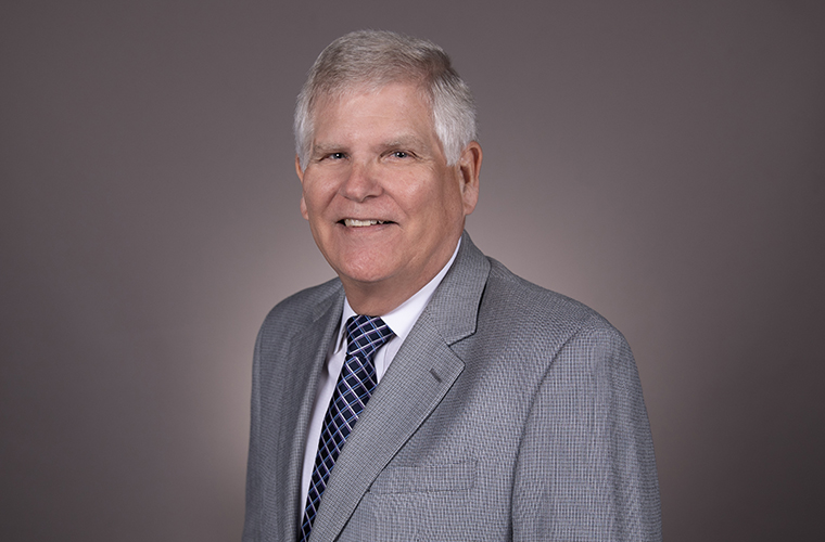 Man with gray hair in a gray jacket, white shirt and blue tie smiling at the camera.