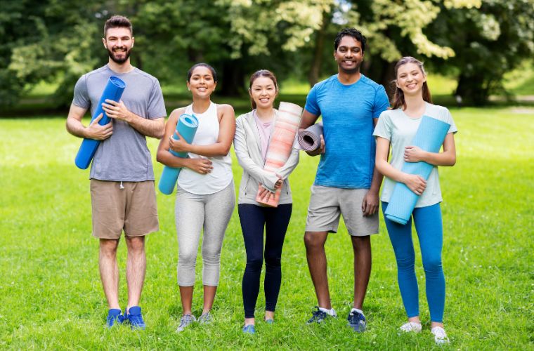 A group of young people, two male and three female, are standing next to each other in athletic attire holding yoga mats and smiling. They are standing in a grassy field with trees in the background.