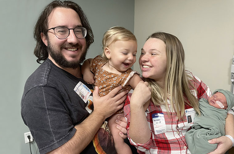 A man smiling at the camera is holding a toddler girl and I woman holds the toddler's hand while also holding a newborn baby.