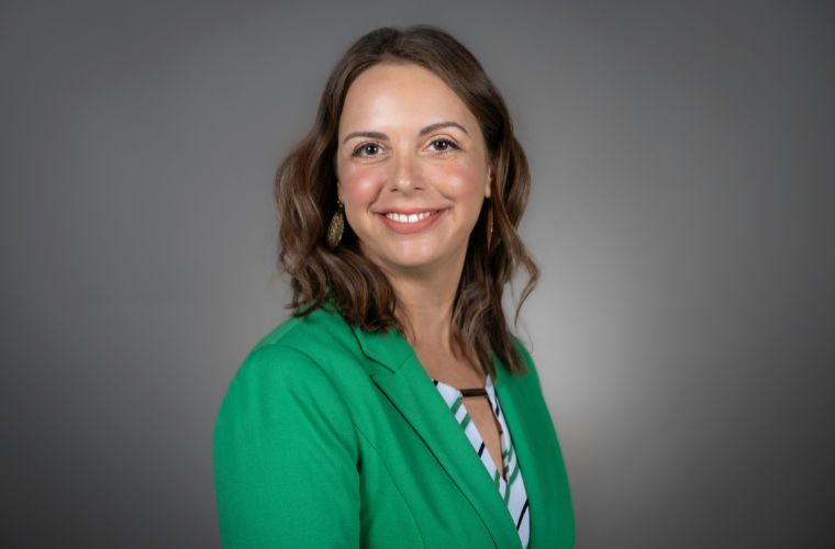 A headshot of Kristy Croom Tucker smiling. She is wearing a green jacket with a green and black striped shirt underneath with round gold earrings. She has shoulder-length wavy light brown hair. 