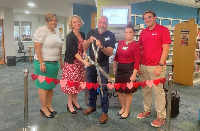 A group of five people are standing lined up in a library at a ribbon cutting event with a gentleman standing in the center holding an oversized pair of scissors.