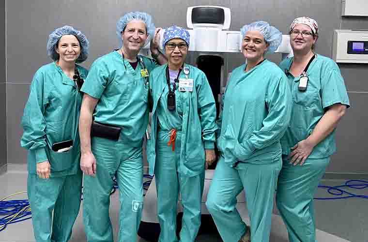 Five members of the surgical team, all wearing green scrubs and surgical caps, stand by the new robotic system.