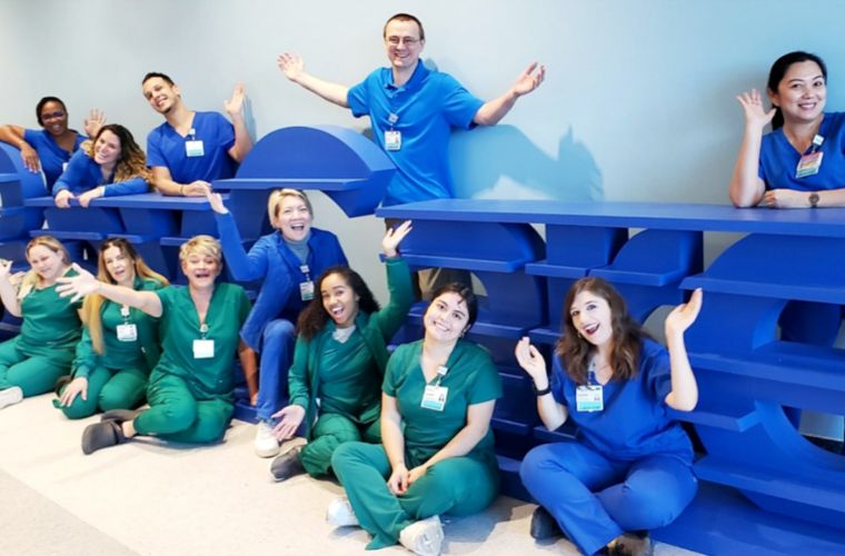 Twelve people wearing scrubs smile widely and gesture enthusiastically as they gather around BayCare sign.