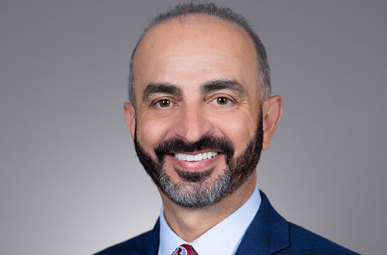 Professional headshot of Dr. Akach. He is wearing a blue suit with white shirt and maroon tie and smiling.