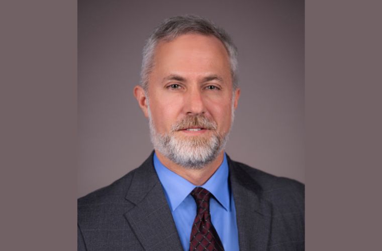An older gentleman with salt and pepper hair and beard smiles in his headshot. He is wearing a blue shirt, a gray jacket and a maroon diamond pattern tie.