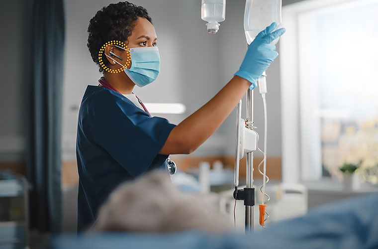 A nurse works at a patient's bedside, wearing a small headset designed to simplify nursing documentation of patient care.