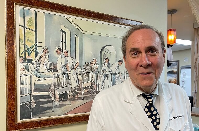 A man in a white coat stands in front of a painting.