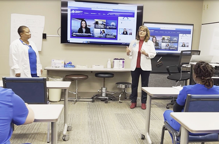 Two women stand in front of a classroom of nursing students.