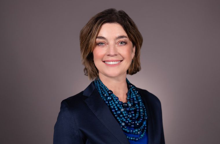 A headshot of Shawn Armstrong smiling. She is wearing a dark navy blazer, a blue shirt and a multi-strand blue beaded necklace. She has chin-length light brown hair parted down the middle.