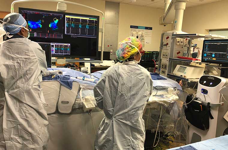 Two clinicians dressed in full white surgical gowns with a blue cap and rainbow tie dye cap respectively, perform the cardiac ablation looking at the console to their right and a big flat screen tv in front of them showing the inside of the heart to guide them with the patient laying on the operating table in front of them.