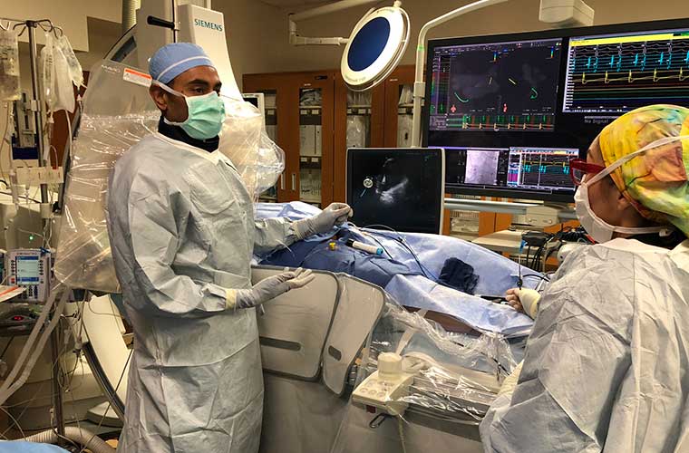  A doctor wearing an all white sterile surgical gown, cap, mask and gloves holds up the POLARx FIT™ Cryoballoon fully inflating inside the operating room with his clinical team. There are screens showing him where to go inside the body of the patient behind him.