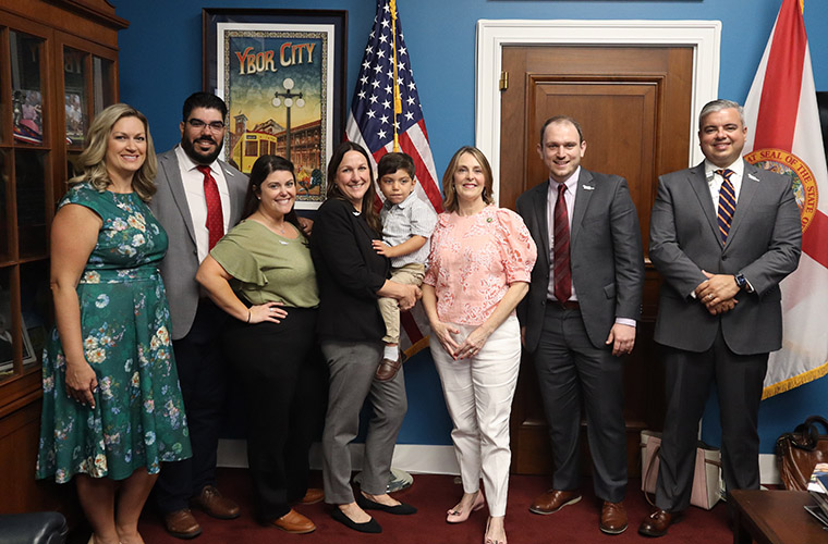 A group of eight people smiling. They are in a bright colored office and woman in middle is holding a toddler.