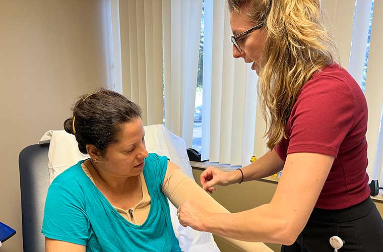 Shelby Green, a lymphedema therapist at the St. Anthony’s Resource Center, is standing wearing a maroon shirt and helps Odalmis Ricardo-Perez slip on a compression sleeve to ease the swelling caused by lymphedema. Ricardo-Perez is seated and wearing a teal short-sleeved shirt.
