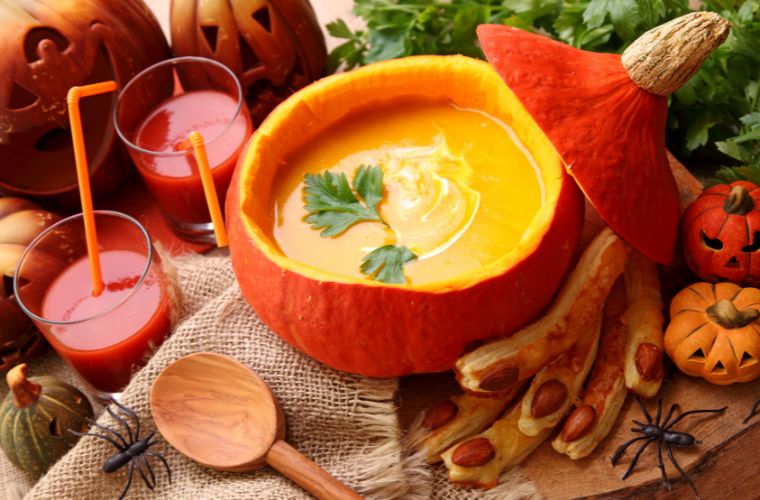 A small bright orange pumpkin with its top cut off resting on the side is filled with pumpkin soup and two sprigs of parsley. It is surrounded by other fall items including carved our pumpkins of various sizes, a wooden spoon, almond pastries, and two glasses of pumpkin juice with orange bendy straws.  In the background is more parsley, and everything is sitting on a wood table covered in a burlap fabric. There are also two black plastic decorative spiders. 