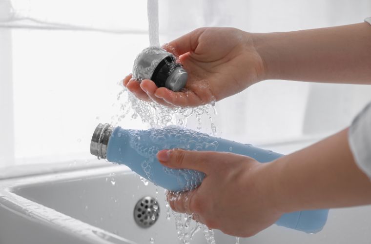 Closeup photo of hands washing a blue water bottle in a white kitchen sink. Water is running down the faucet as it rinses the bottle's cap and exterior of the bottle.