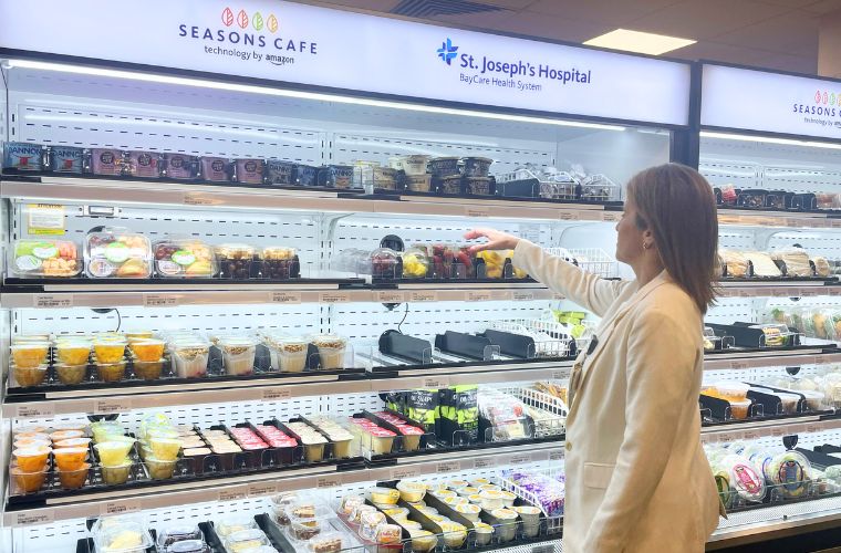 A woman with short blonde hair wearing a white blazer reaches for a food item in a cafeteria. There is signage that reads SEASONS CAFE technology by amazon and St. Joseph's Hospital BayCare Health System above the cooler.