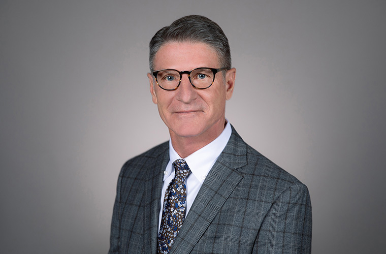 A person dressed in a grey suit poses for a professional photo in front of a grey backdrop.