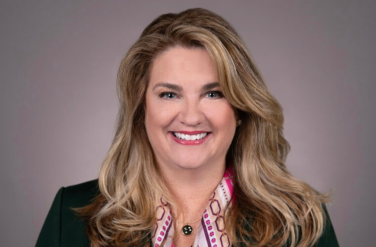 A smiling headshot of Jennifer McVan. She is wearing a black jacket with a patterned shirt underneath and a round silver necklace. Her long blonde wavy hair is parted on the side.
