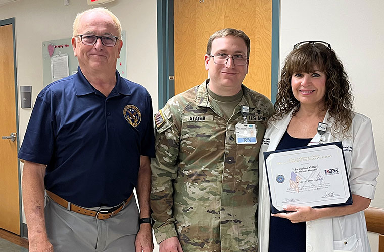 A woman in a white medical coat holds up a certificate as she stands with two men, one dressed in a polo and slacks and the other dressed in military fatigues.