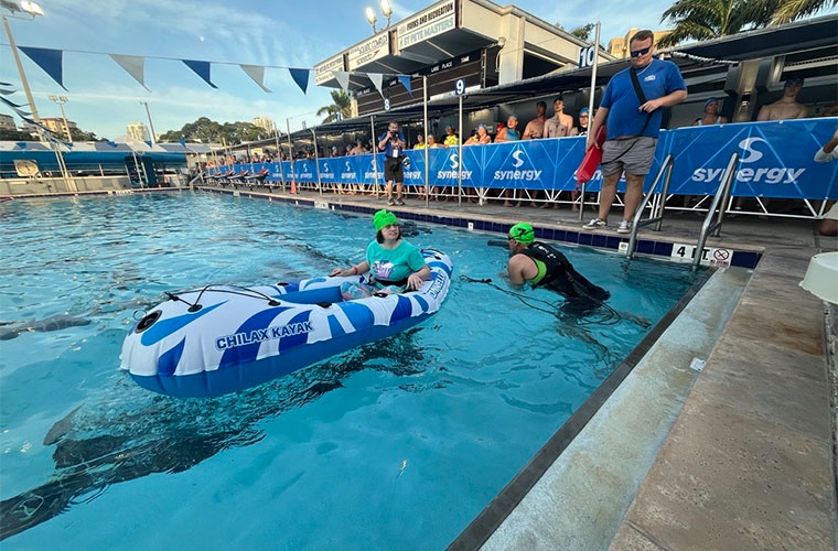 A man wearing a swimsuit tows an inflatable boat carrying a young woman in a pool during an athletic competition.