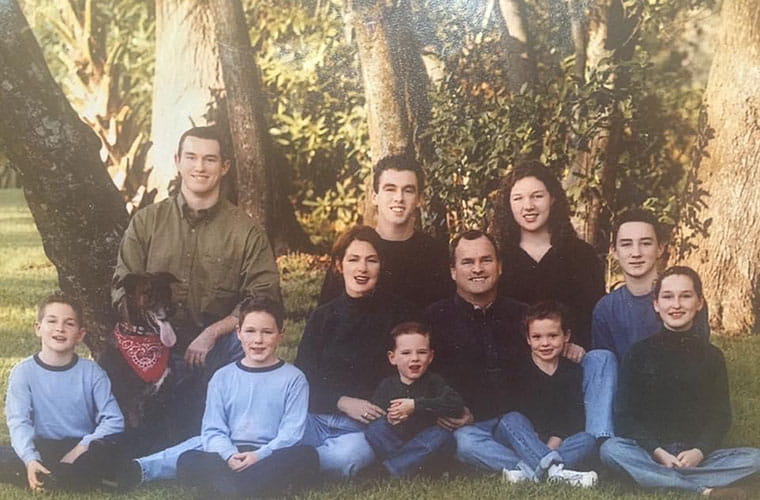 A large group with a man and women and several boys and girls dressed in similar clothing sit in the grass with a dog for a family portrait.