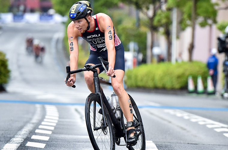 A young man in an athletic suit and bicycle helmet rides a bike on the street in a race.