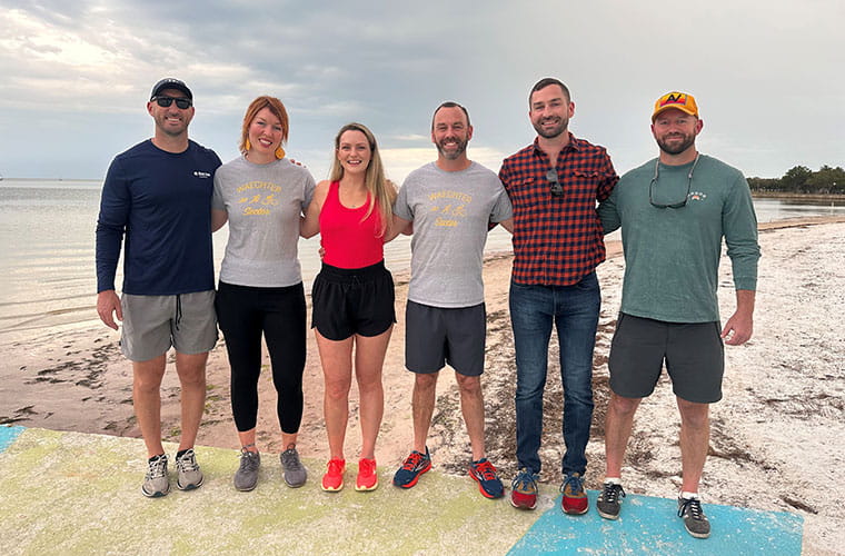 Four men and two women stand arm and arm along a beach.