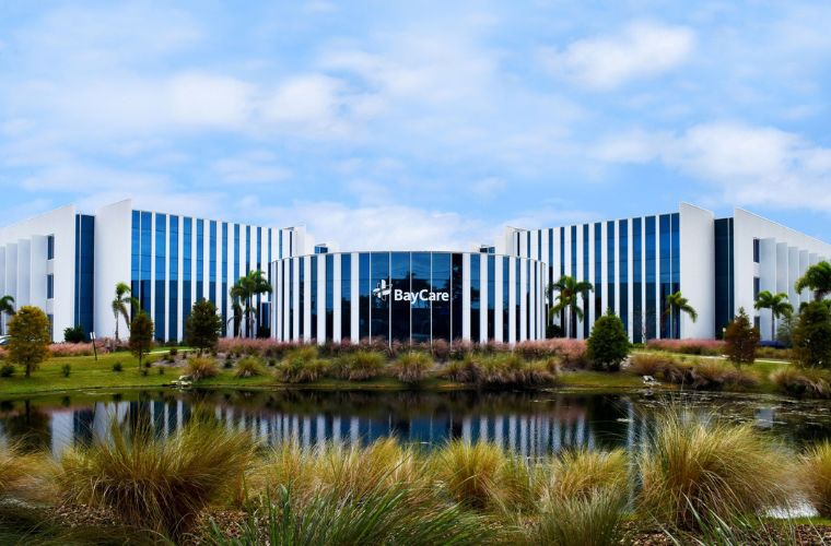 A modern office building is encircled by lush, tall grass, blending with a lake in front of it. The word BayCare is displayed on the front of the building. 