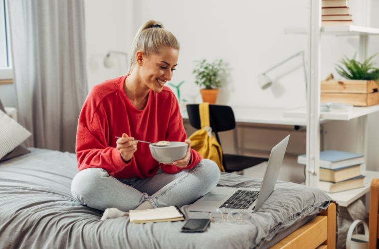 A woman sits on her bed in a dorm room, using a laptop while enjoying a bowl of cereal.