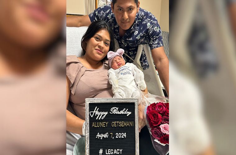 New parents smiling in a hospital bed holding a newborn baby in a white onesie that reads 'I was born at the legacy hospital.'