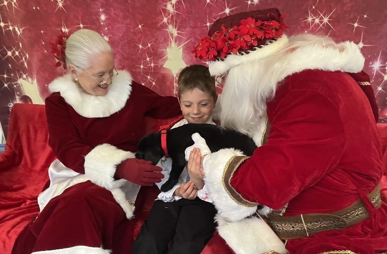 A young boy holds a Black Labrador Retriever puppy with a red collar. He is seated between Santa Claus and Mrs. Claus dressed in red costumes in front of a festive background.