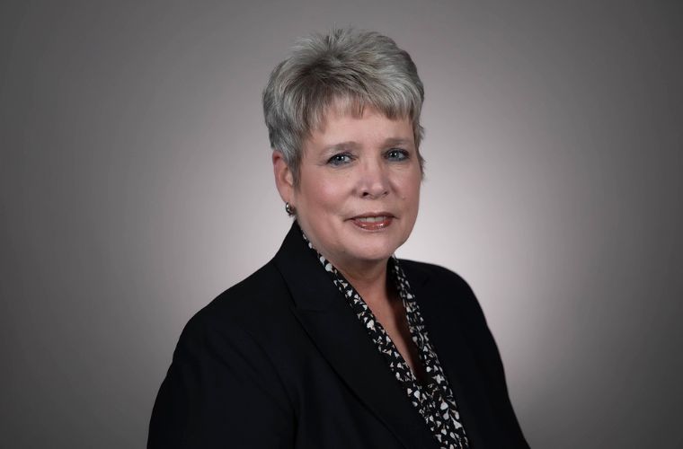 A headshot of Donna Lynch smiling. She is wearing a black jacket with a black and white floral print shirt underneath with small silver hoop earrings. She has very short, spiky salt and pepper color hair. 