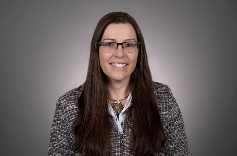 A headshot of Danielle Mauck smiling. She is wearing a tweed-style jacket in shades of brown with a white collared shirt underneath. She is wearing glasses and has on a large round pendant silver necklace. Her long straight brown hair is parted down the middle.